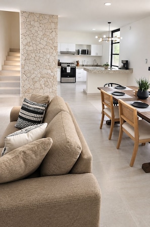 Un salon moderne avec un canapé beige, un coin repas avec des chaises en bois, une cuisine avec des meubles blancs et un mur en pierre.