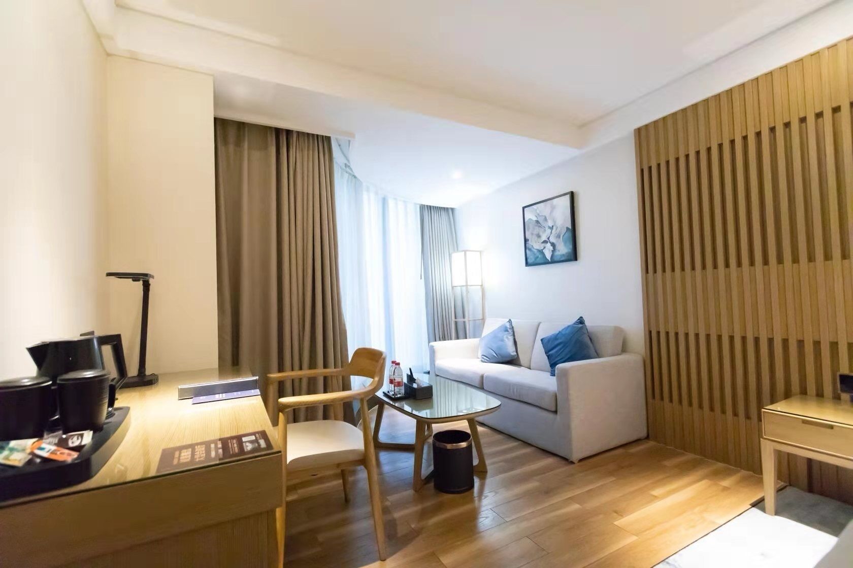 Habitación de hotel moderna con suelo de madera, sofá blanco con cojines azules, una mesa de centro de vidrio, una silla de madera, un escritorio con computadora y una lámpara, y un ventanal con cortinas.