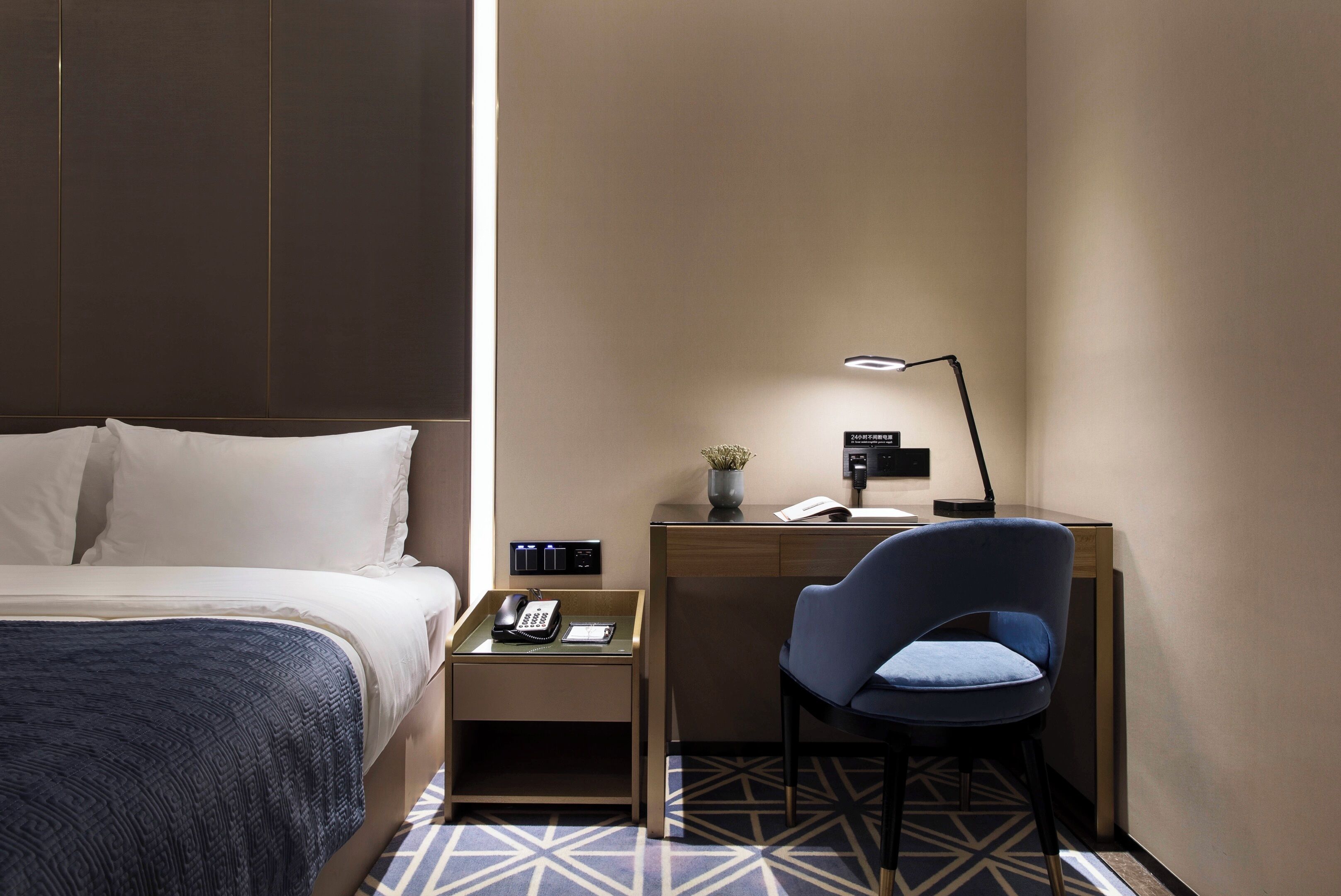 Habitación de hotel con cama, escritorio con silla, lámpara de escritorio, teléfono y una pequeña planta en maceta.