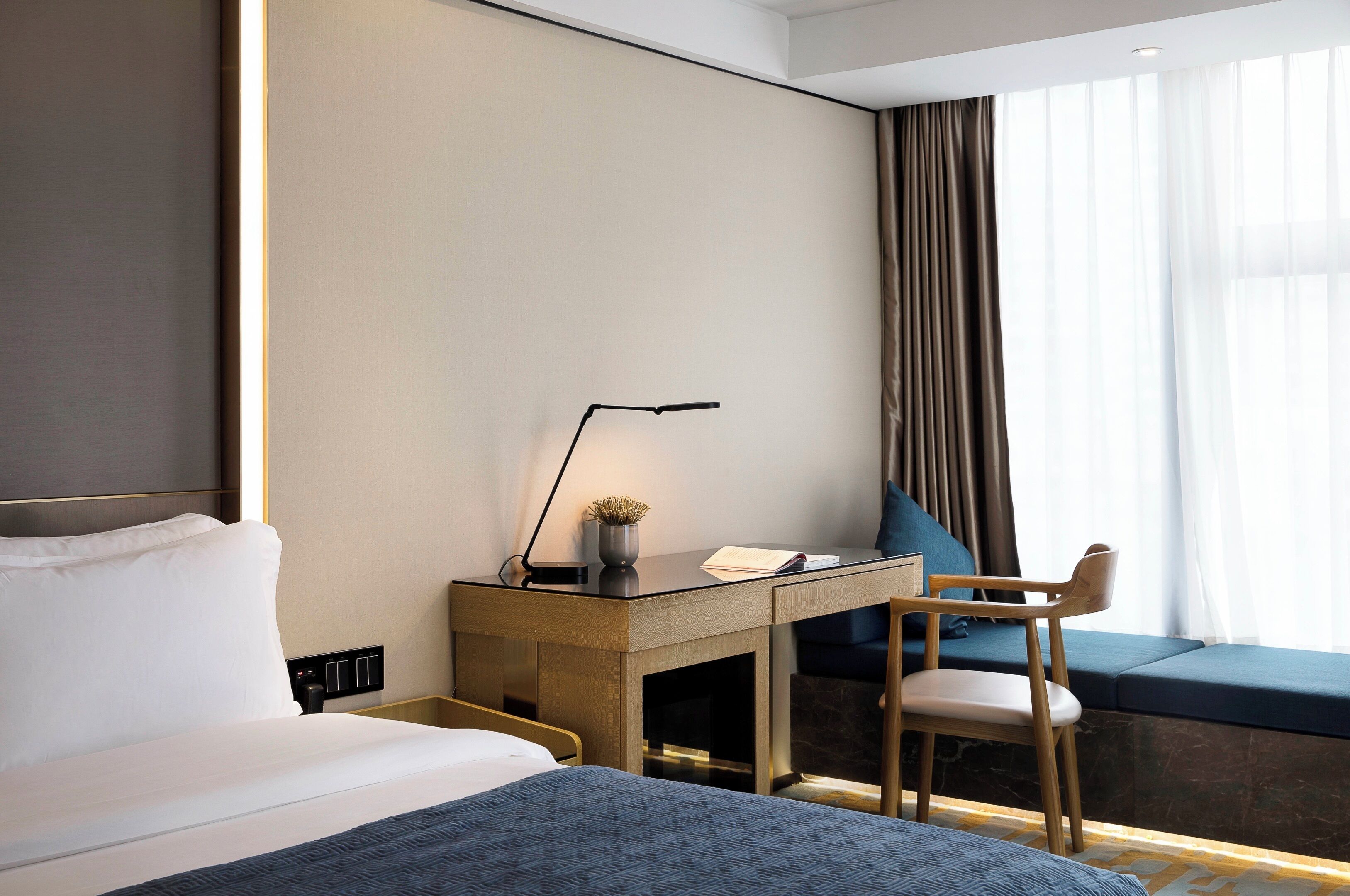 A hotel room with a bed, a desk with a lamp, a chair, and a window with curtains.