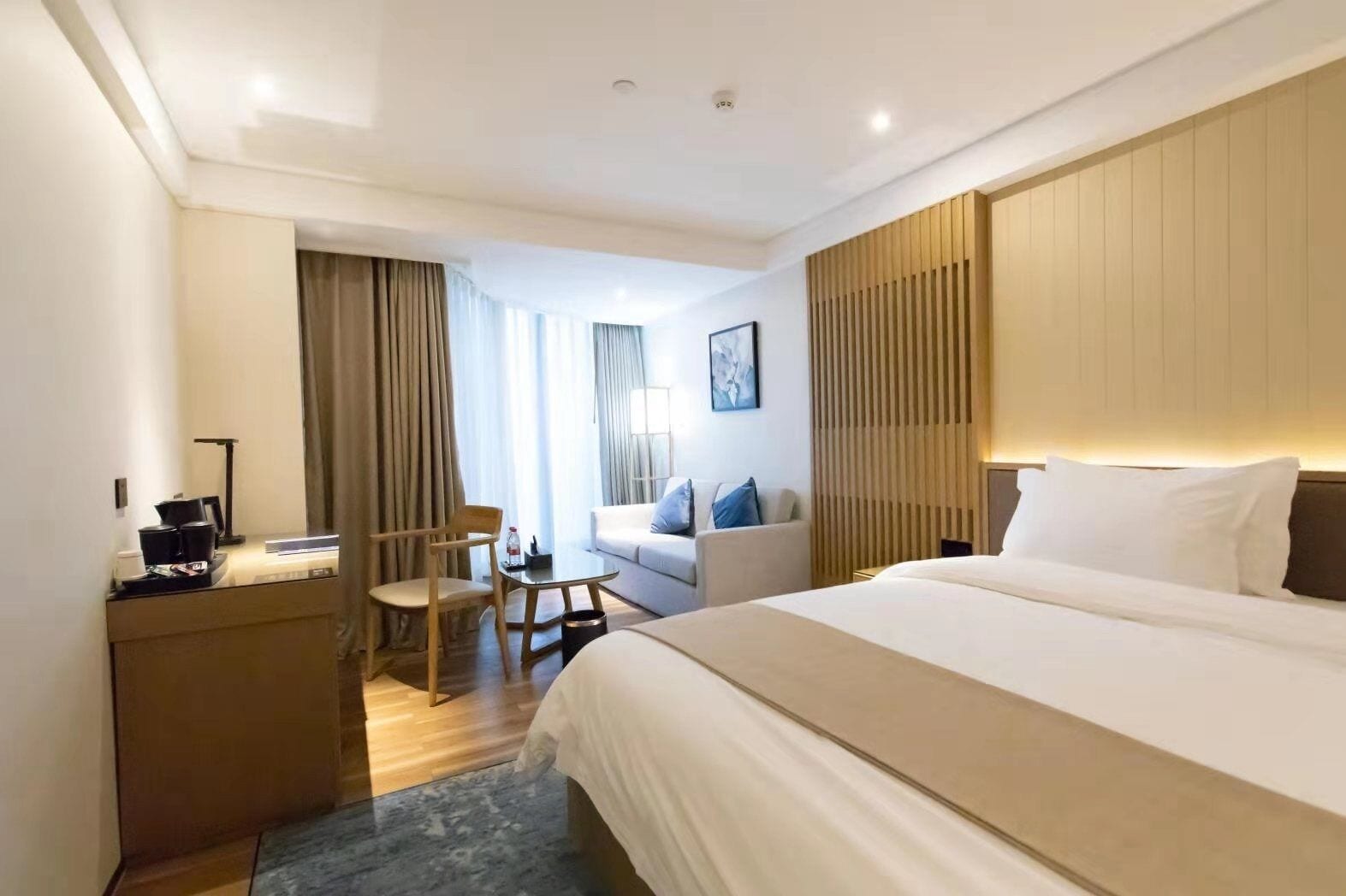 A hotel room with a bed, a desk, a chair, a sofa, and a window with curtains.