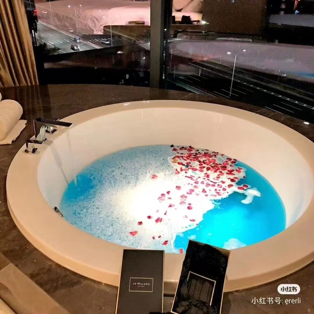 A large bathtub filled with water and rose petals, with a view of a cityscape through a window.