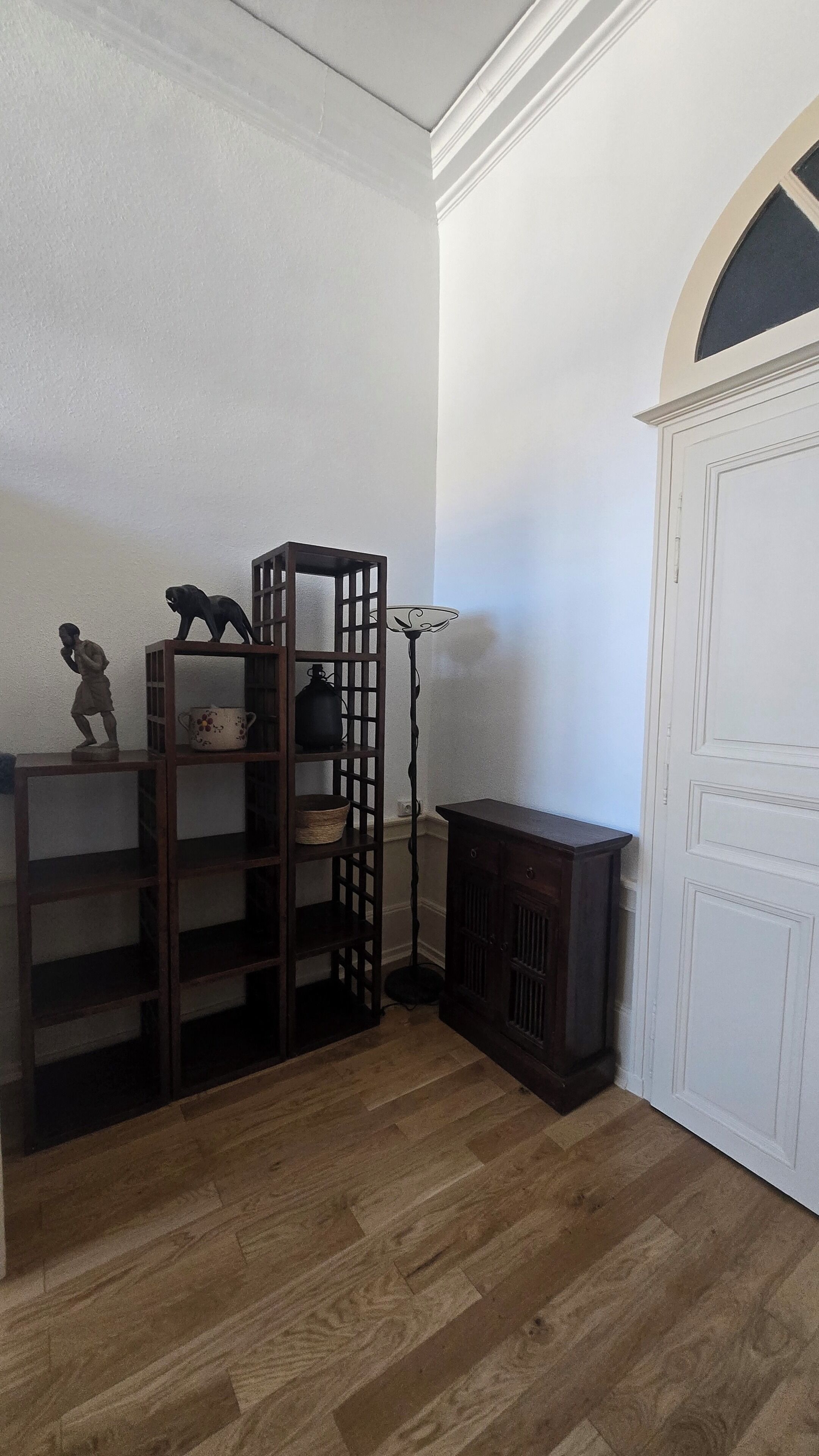 Una habitación de esquina con piso de madera, un mueble de madera oscura, una puerta blanca con arco decorativo y un estante con adornos.
