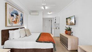 A bedroom with a bed, a TV, a painting, and a kitchenette in the background. - Port Douglas Studio with Balcony Spa Bath | Ivy Pearl Studio (Port Douglas)