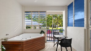 A bathroom with a wooden bathtub, a small table with chairs, and a view of a garden through a window. - Port Douglas Studio with Balcony Spa Bath | Ivy Pearl Studio (Port Douglas)