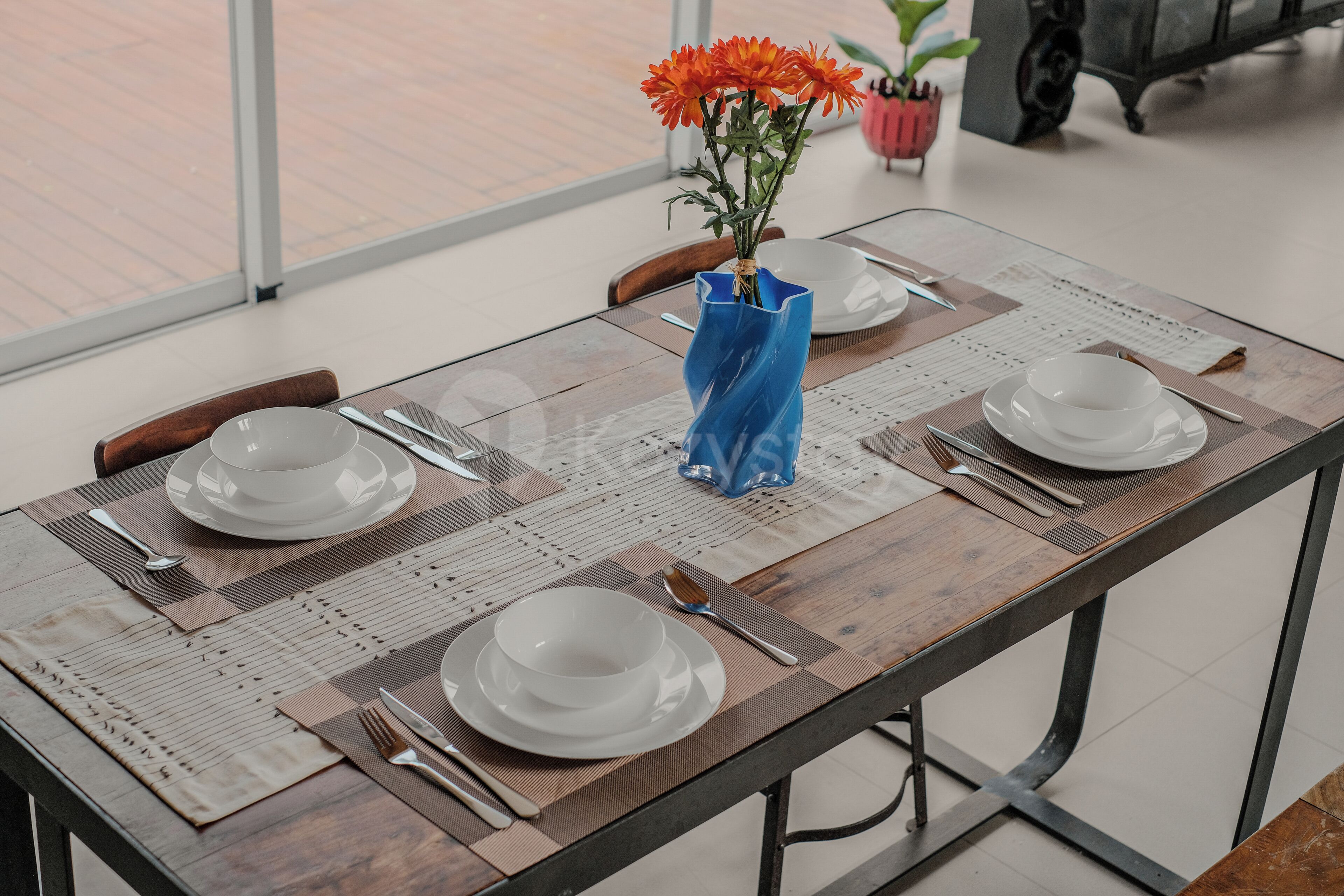 A dining table set with white plates, silverware, and a blue vase with orange flowers.