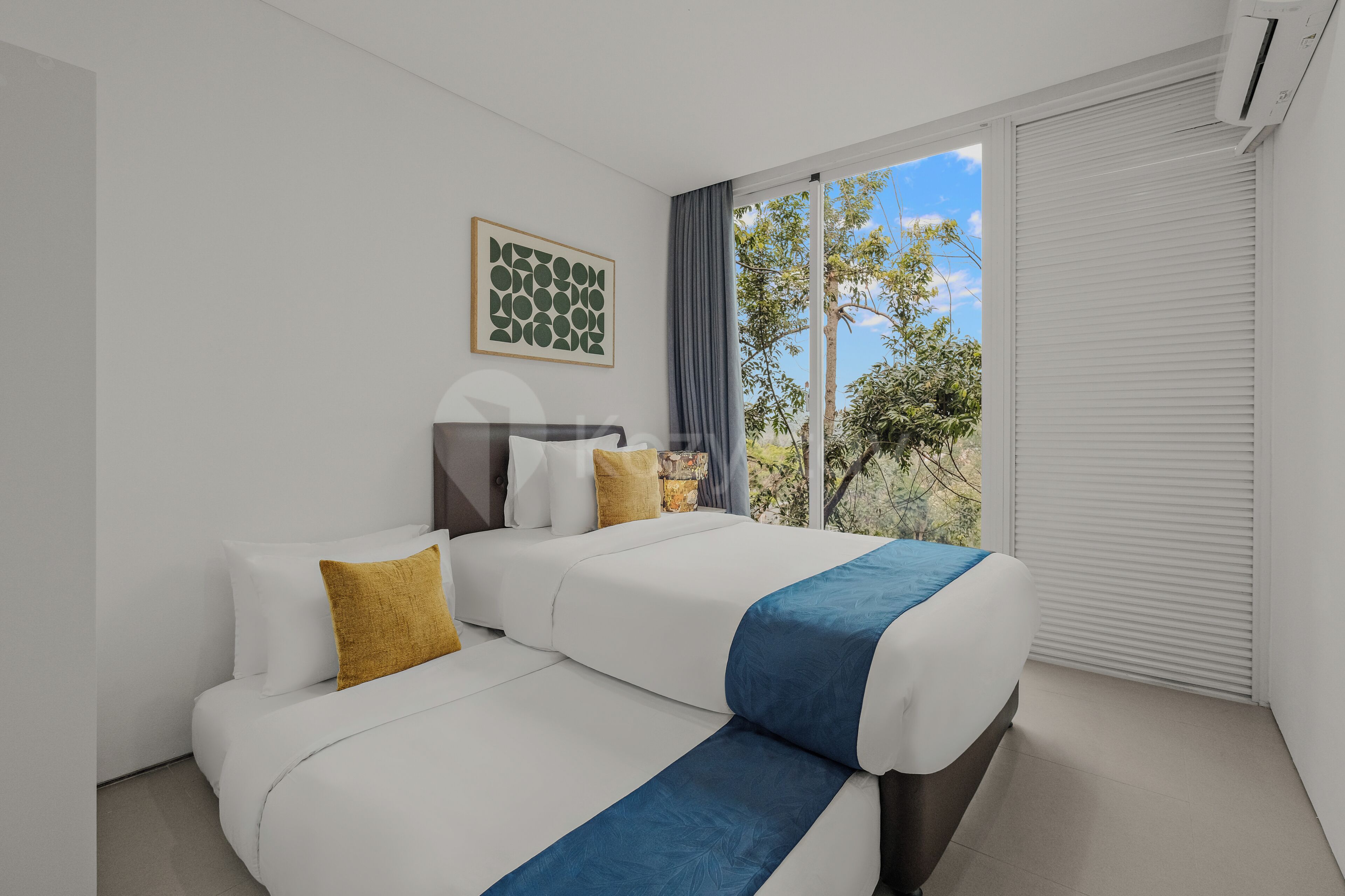 A modern hotel room with a bed, two bedside tables, a window with a view of trees, and a wall-mounted artwork.