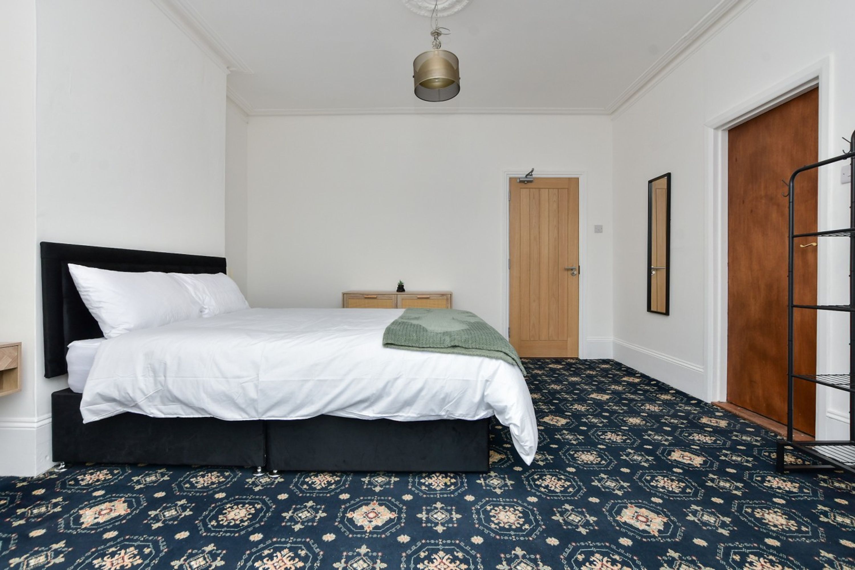 A bedroom with a bed, a nightstand, a wooden door, and a patterned carpet.