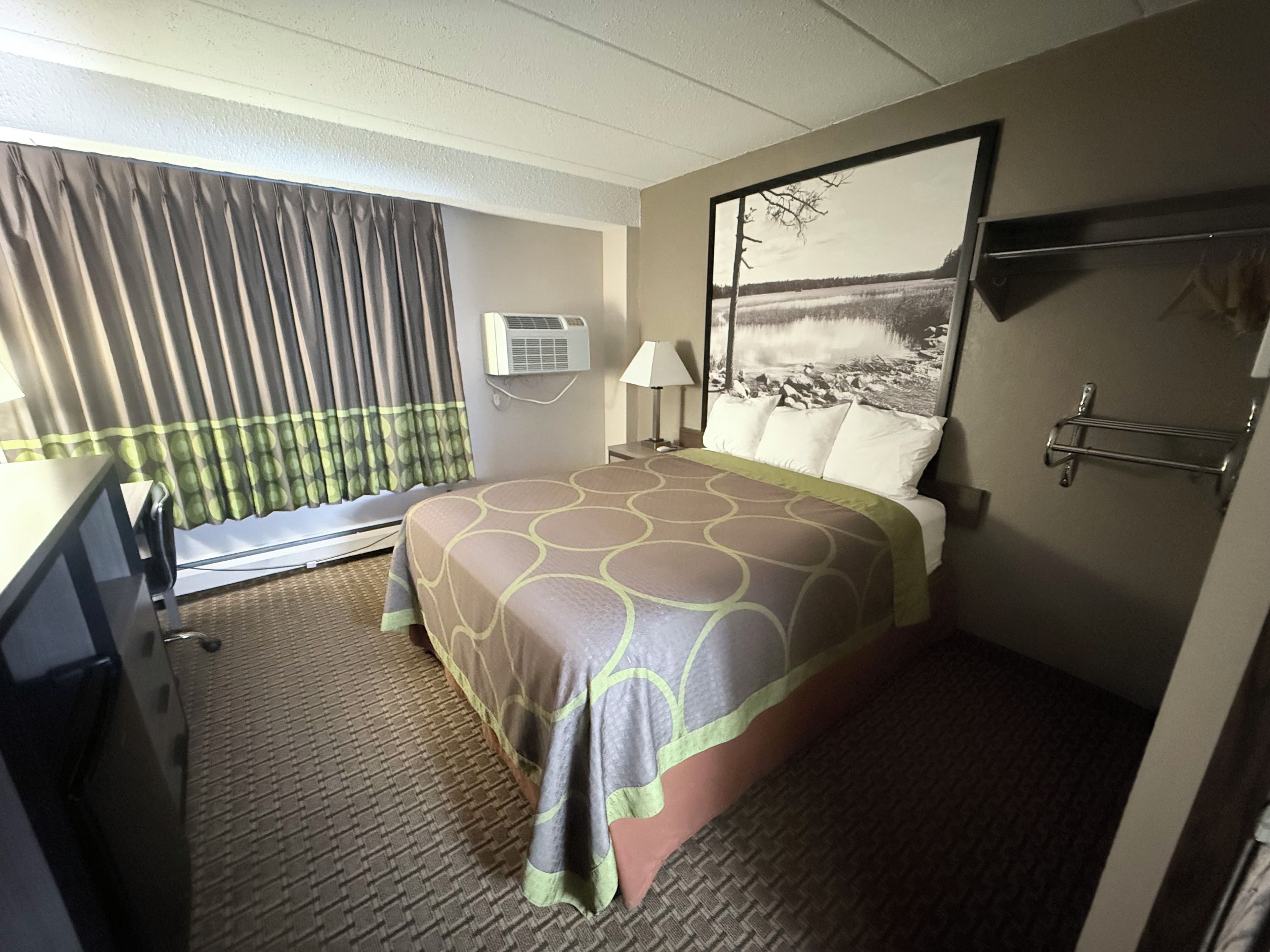 A hotel room with a bed, a desk, a chair, a window with curtains, an air conditioning unit, a lamp, and a painting on the wall.