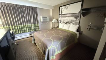 Habitación de hotel con cama, escritorio, silla, ventana con cortinas, equipo de aire acondicionado, lámpara y un cuadro en la pared.