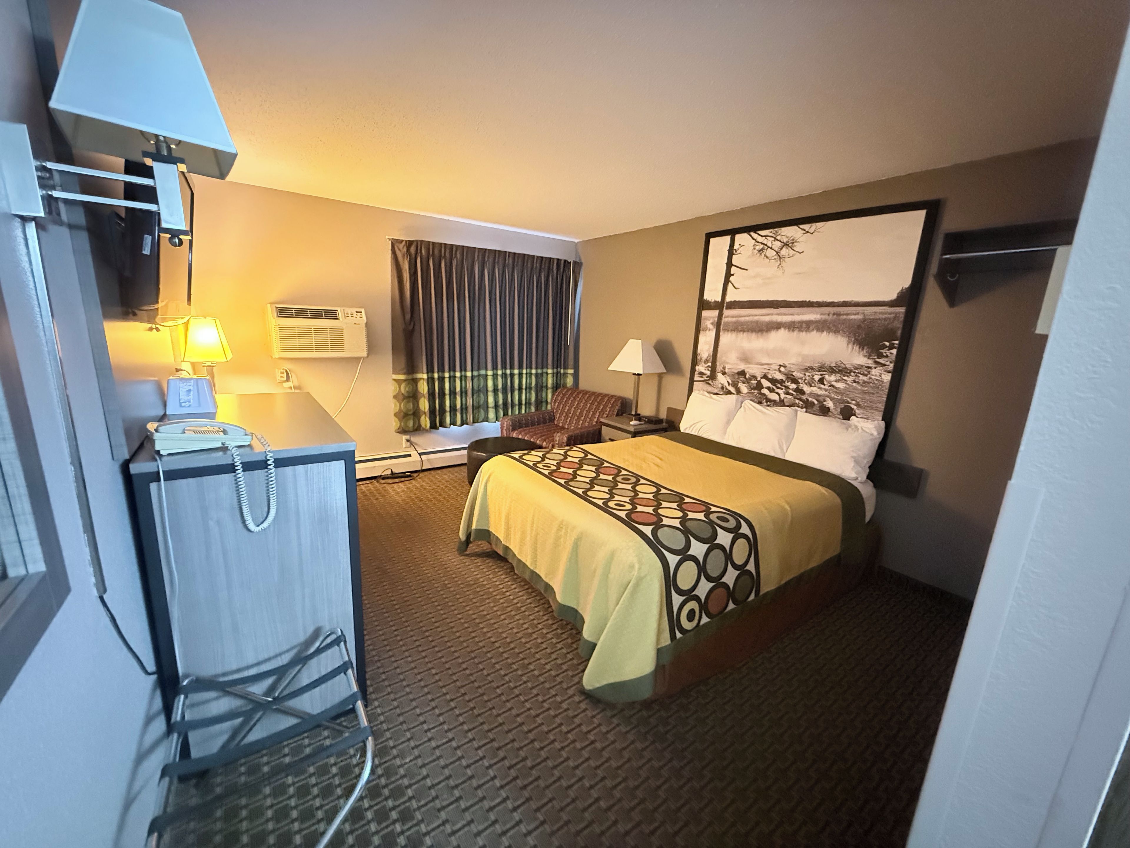 Quarto de hotel com cama, escrivaninha com telefone, luminária, janela com cortinas e um quadro na parede.