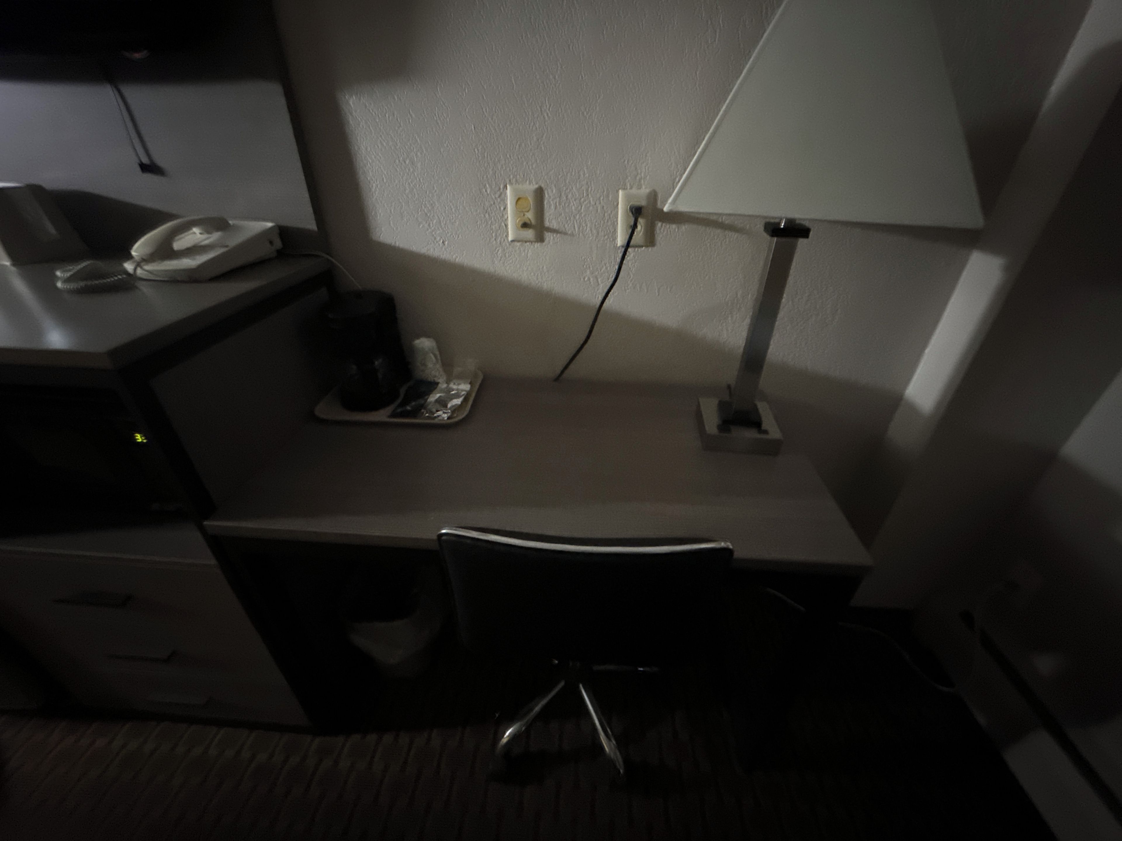A hotel room with a desk, a chair, a lamp, a phone, and a coffee maker.