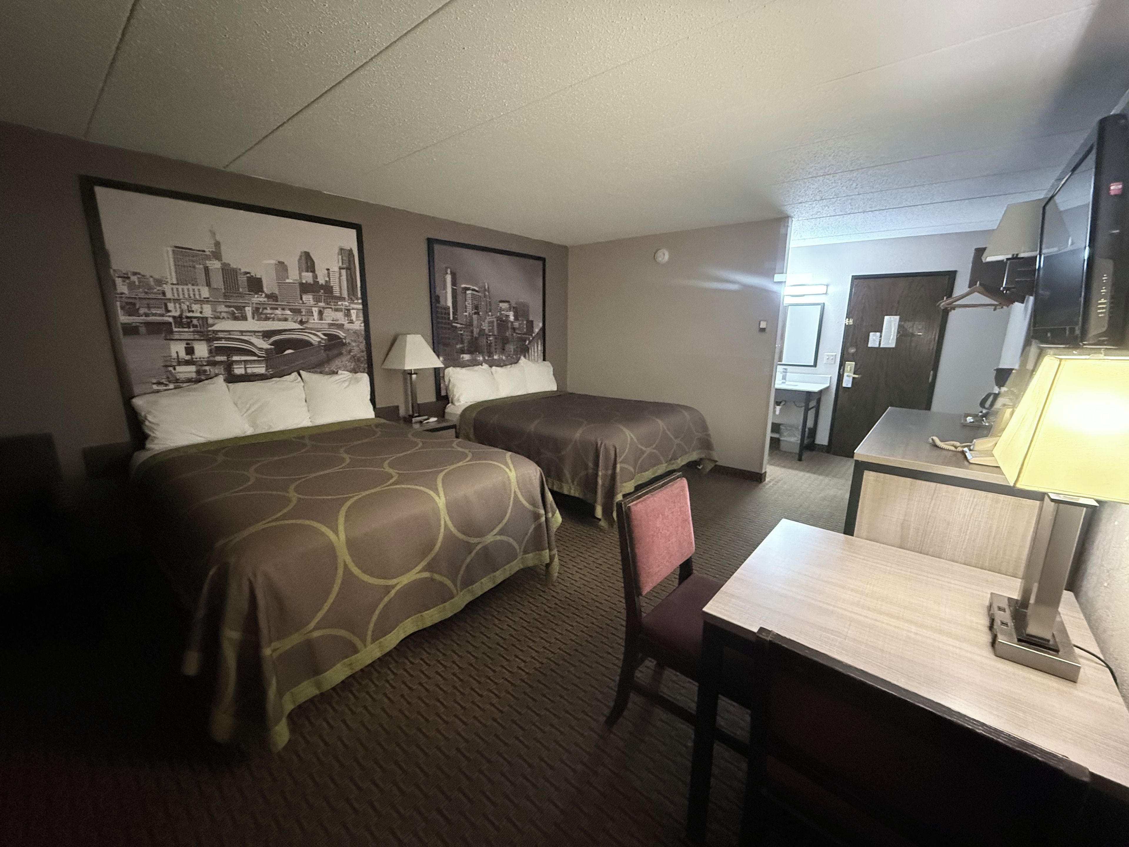 A hotel room with two beds, a desk, a chair, a lamp, a television, and a cityscape poster on the wall.