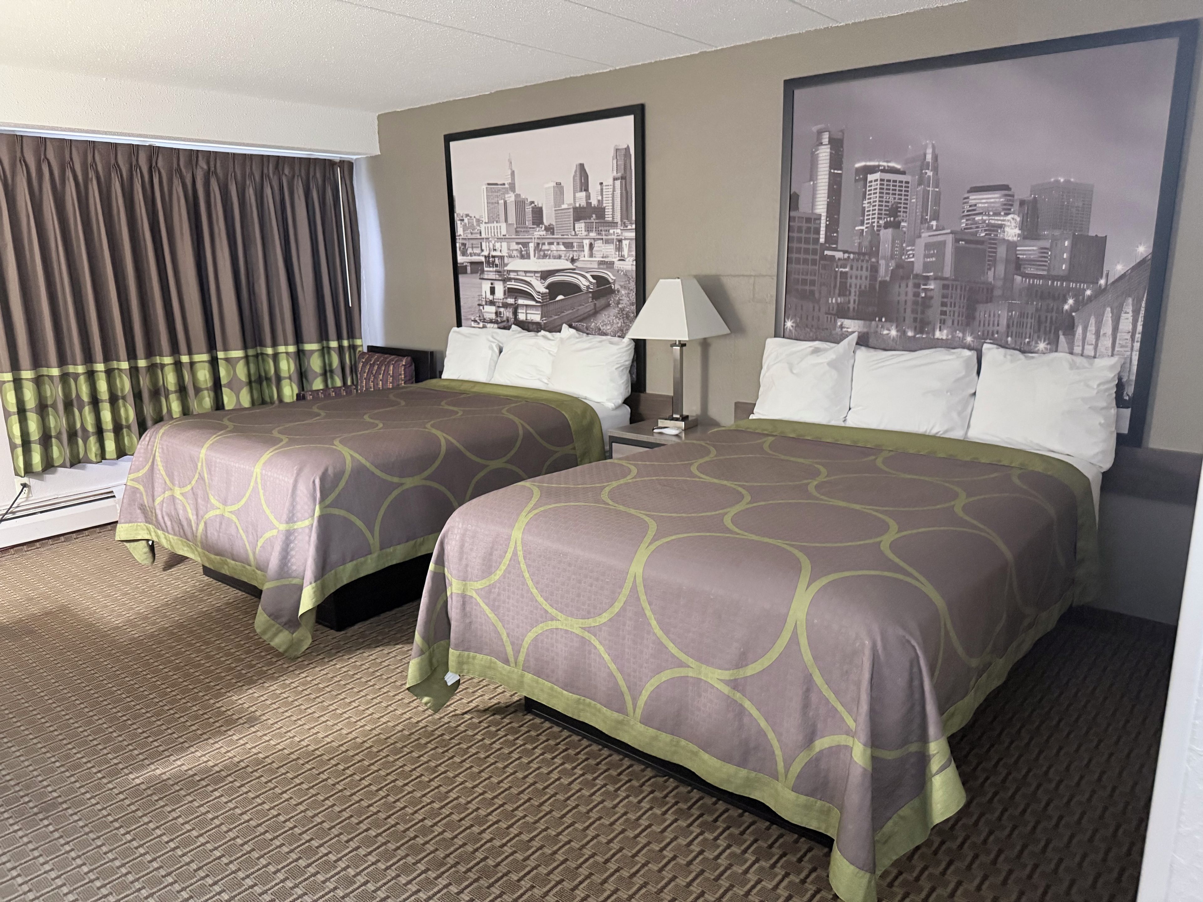 A hotel room with two beds, a window with curtains, a lamp, and two framed cityscape pictures on the wall.