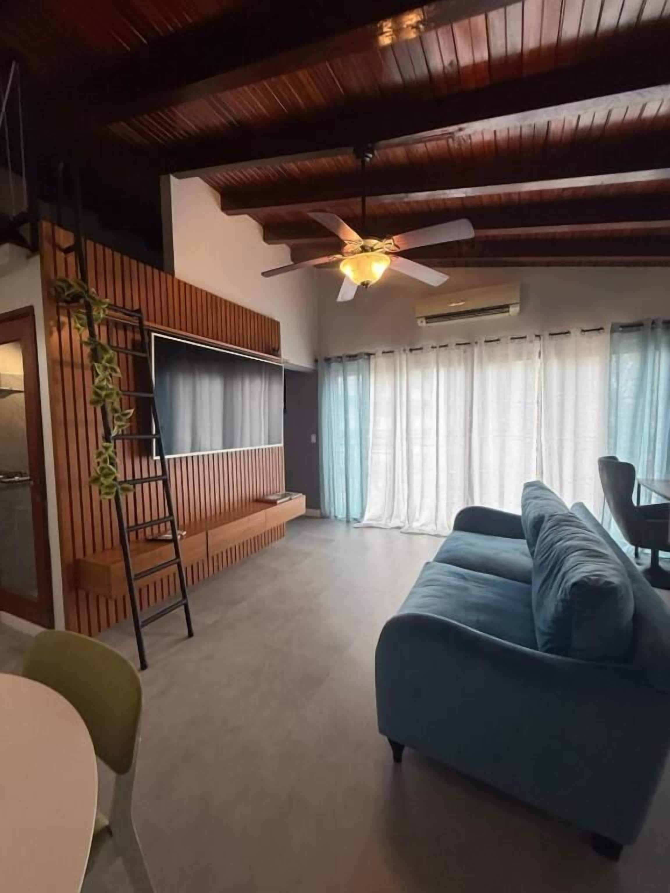 Sala de estar com um sofá azul, uma escada de madeira, um ventilador de teto e uma janela com cortinas.