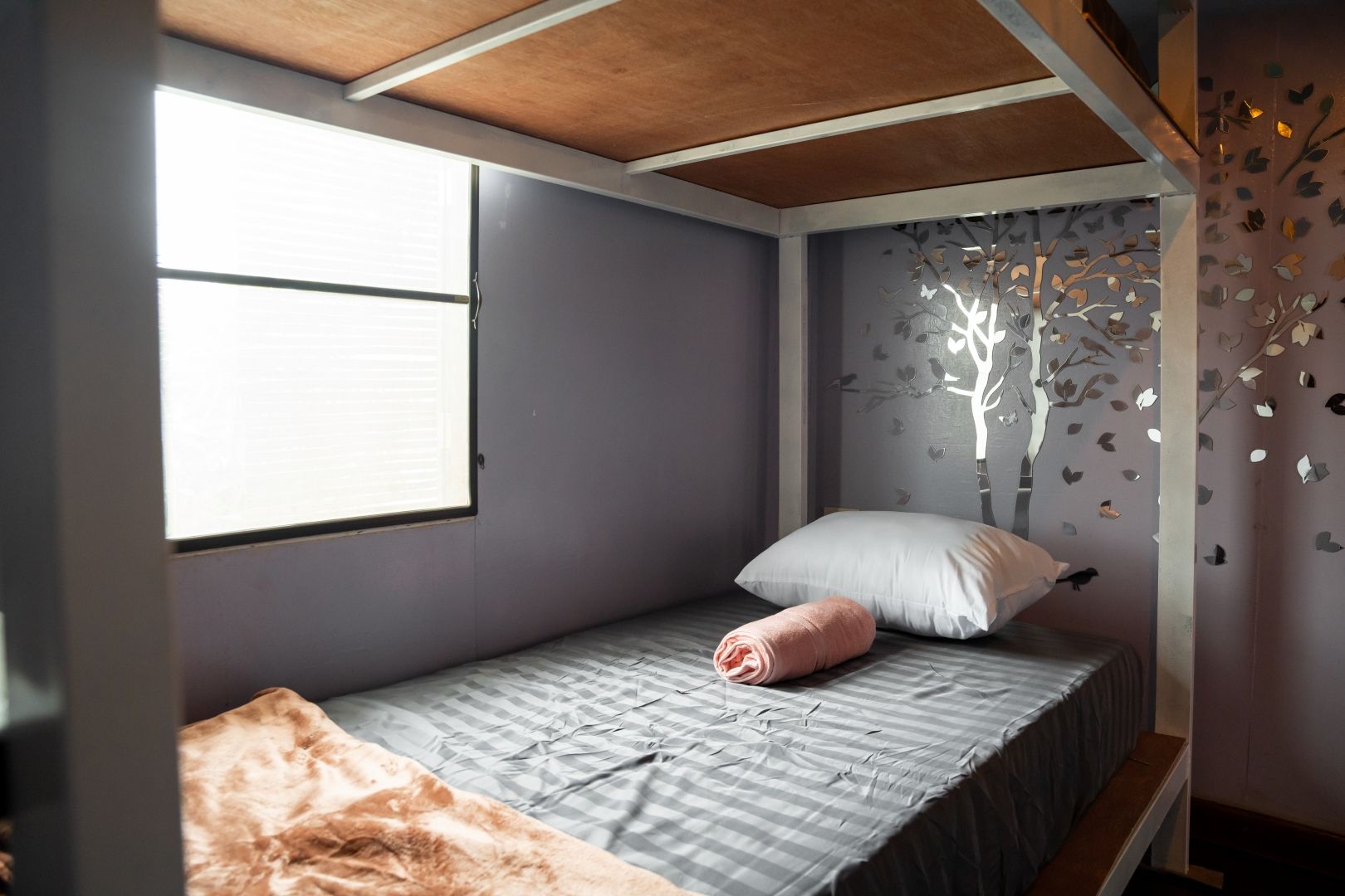 Una cama individual con colcha gris a rayas, una almohada blanca y una toalla rosa. Hay una ventana con persianas, una pared con un adhesivo de árbol y un techo de madera.