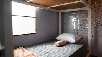 Una cama individual con colcha gris a rayas, una almohada blanca y una toalla rosa. Hay una ventana con persianas, una pared con un adhesivo de árbol y un techo de madera.