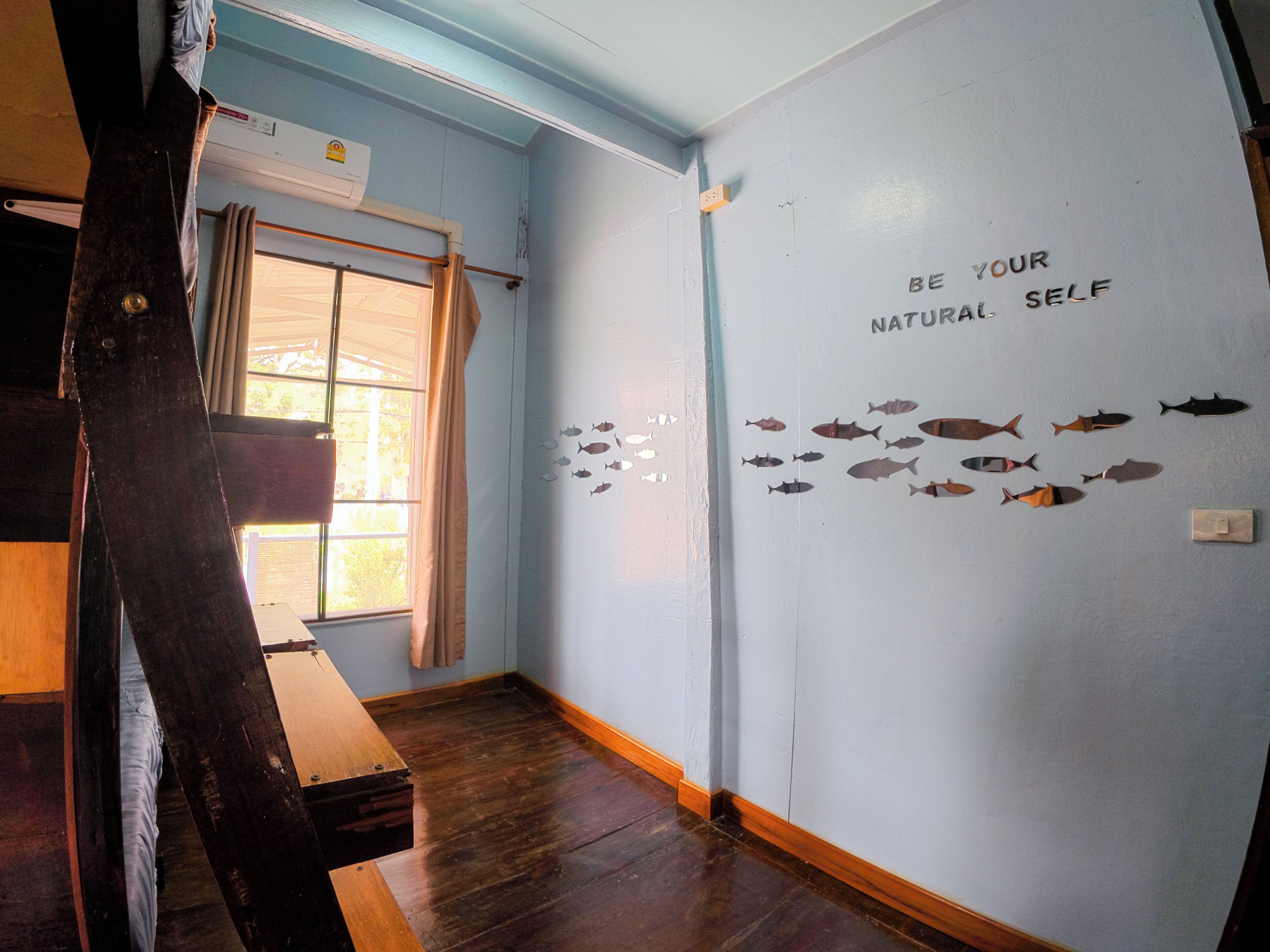 A room with a wooden floor, a window with curtains, and a wall with fish silhouettes and the text 'BE YOUR NATURAL SELF'.