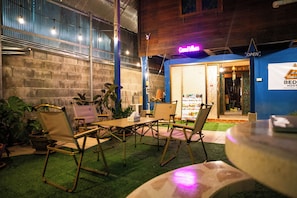 An outdoor seating area with tables and chairs, a neon sign, and a building entrance with a sign.