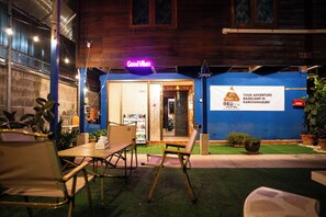 An outdoor seating area with tables and chairs, a neon sign reading 'Good Vibes', and a sign for 'Bed 4U Hostel' advertising a basecamp in Kanchanaburi.