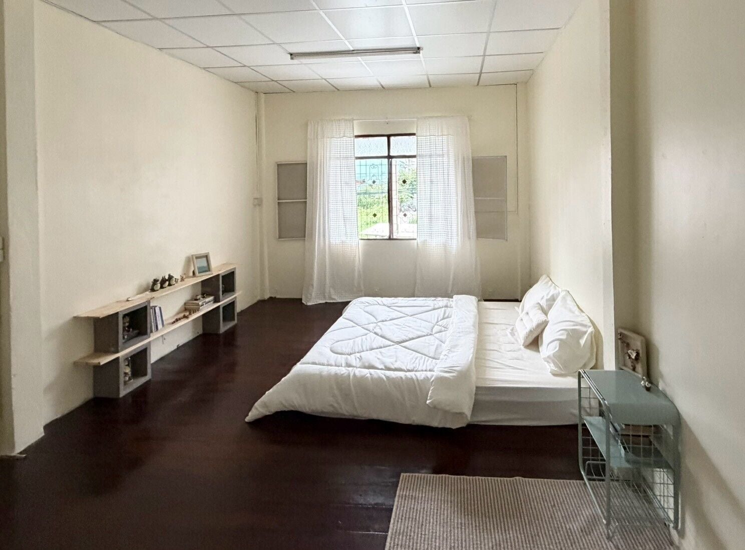 A bedroom with a bed, a window with curtains, a shelf with items, and a radiator.