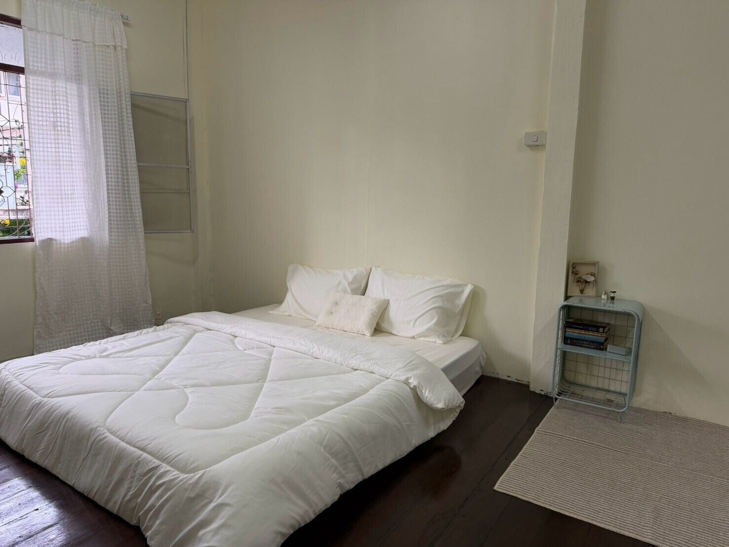 A bedroom with a bed, white bedding, a window with a curtain, a bedside table with books, and a small shelf.
