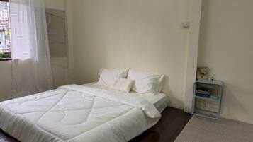 A bedroom with a bed, white bedding, a window with a curtain, a bedside table with books, and a small shelf.