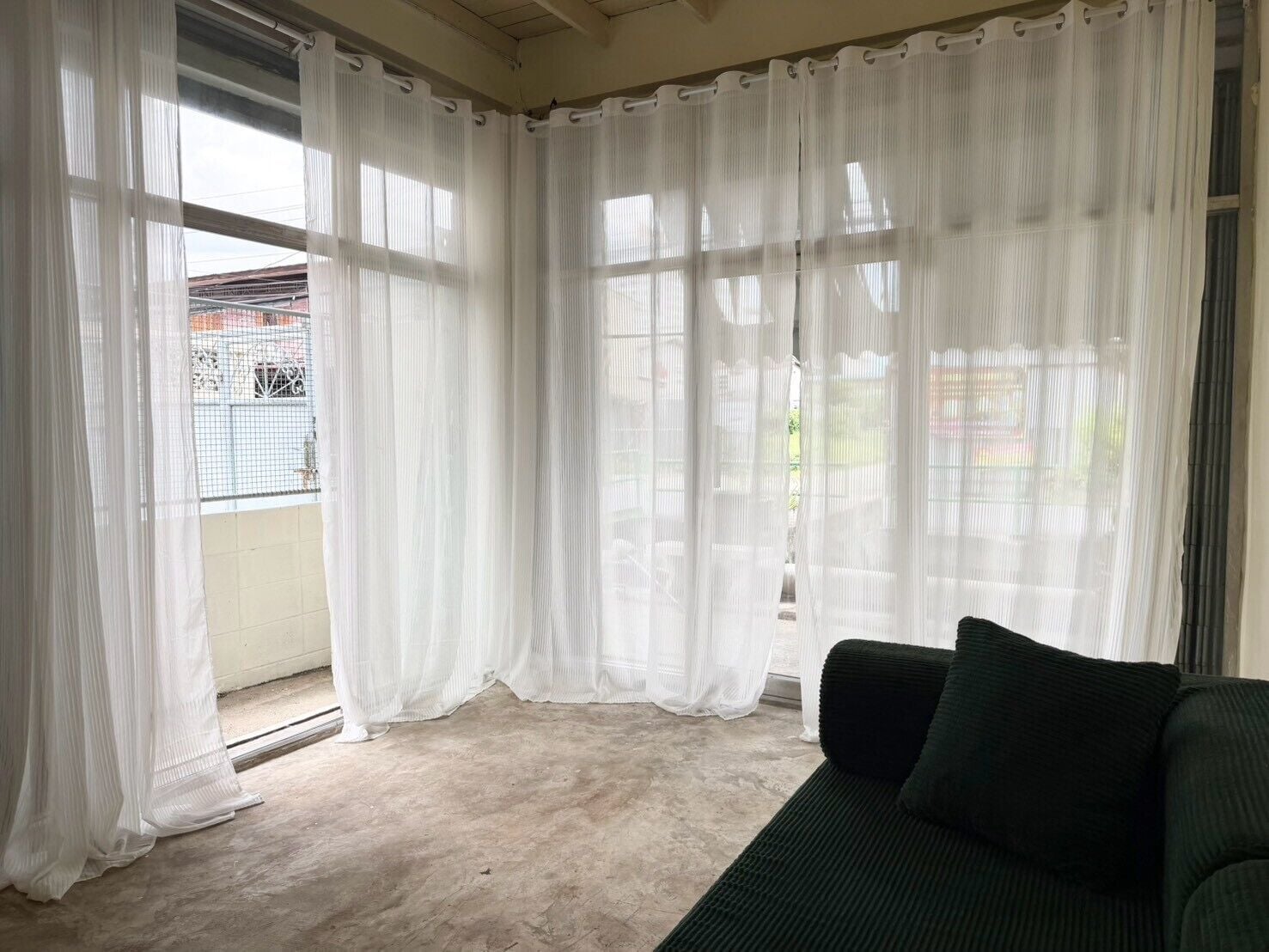 A room with a green sofa, white curtains, and a view of a balcony with a metal railing.