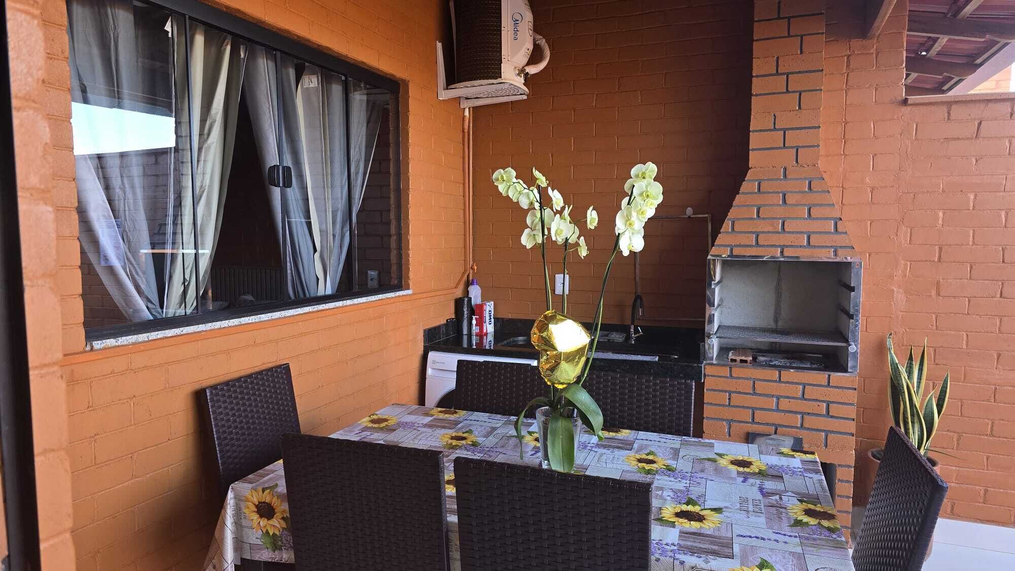 Een terras buiten met een bakstenen muur, een tafel met een bloemen tafelkleed, stoelen, een barbecue en een raam met gordijnen.