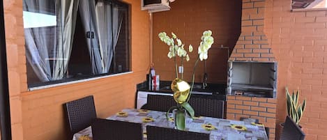 An outdoor patio with a brick wall, a table with a floral tablecloth, chairs, a grill, and a window with curtains.