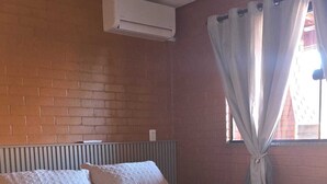 Ein Zimmer mit einem Einzelbett, einer Ziegelwand, einem Fenster mit Vorhängen und einer Klimaanlage.