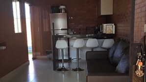 Eine moderne Küche mit Kühlschrank, Mikrowelle und Kaffeemaschine. Ein Essbereich mit Tisch und Stühlen ist vorhanden. Der Raum verfügt über ein großes Fenster und eine Couch.
