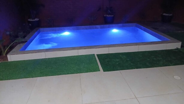 Ein rechteckiger Pool mit blau beleuchteter Wasseroberfläche, umgeben von einer gefliesten Liegefläche und grüner Kunstrasen.