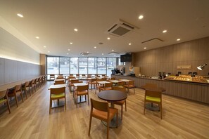 A spacious indoor area with multiple wooden tables and chairs, a counter with food display, and large windows providing natural light. - R B Hotel Sendai East (Sendai)
