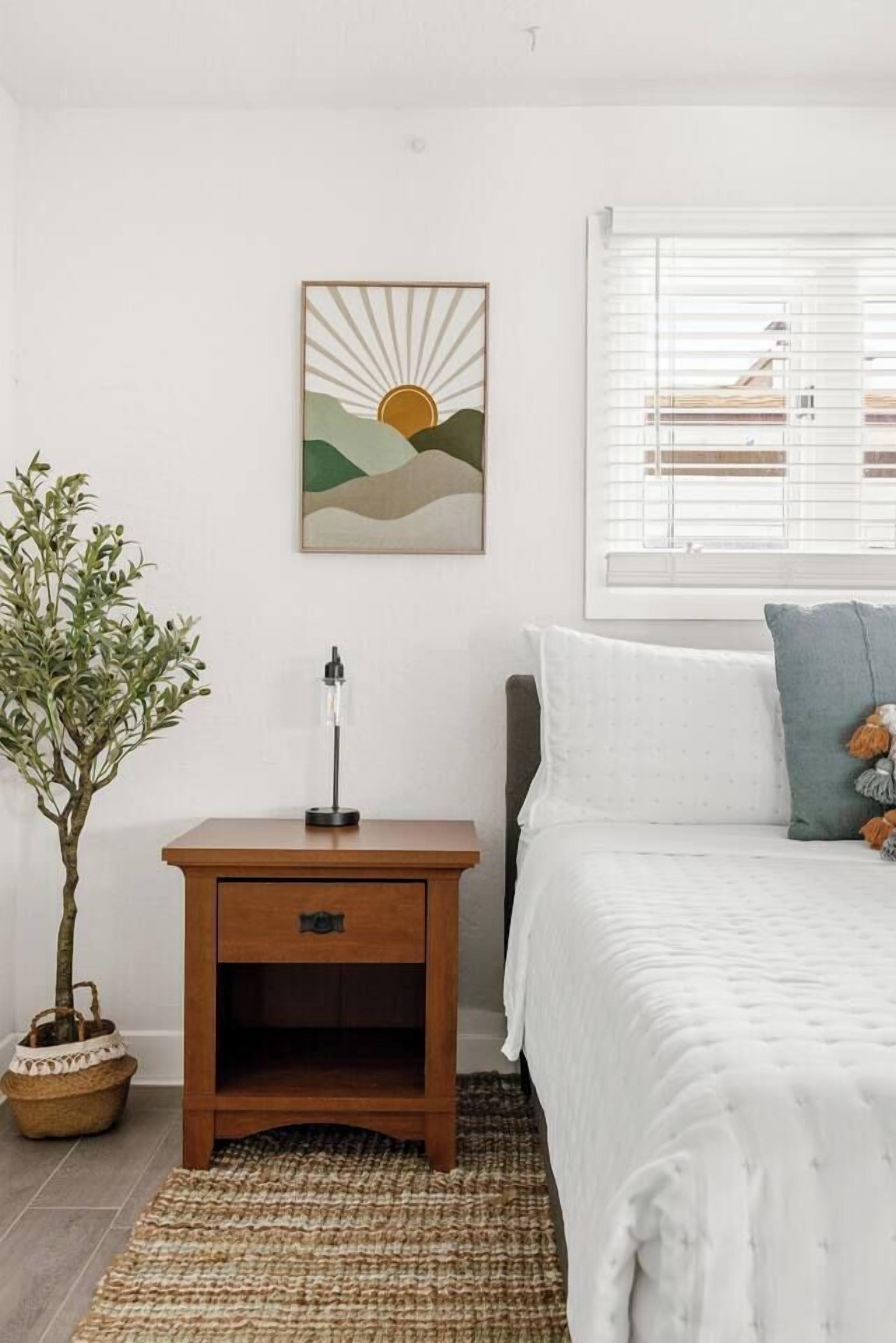 Un dormitorio con cama, mesita de noche, lámpara, un cuadro en la pared, ventana con persianas y una planta en maceta.