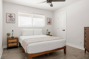 Ein Schlafzimmer mit Bett, Nachttischen, Deckenventilator, Fenster mit Jalousien und Bildern an den Wänden.