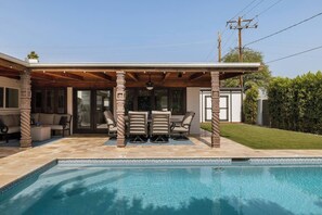 A swimming pool with a covered patio area, outdoor furniture, and a view of the backyard.