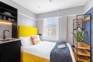 Una habitación de hotel moderna con cama, mesita de noche y ventana con vistas a edificios.