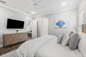 Quarto com cama, cômoda, televisão de tela plana, ventilador de teto e porta que dá para outro espaço.