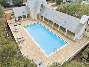 Uma piscina retangular com um deck ladrilhado, rodeada por espreguiçadeiras e uma área de varanda coberta.