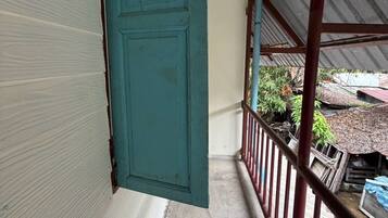 A narrow corridor with a teal door, a metal railing, and a view of a yard with plants and a building in the background.