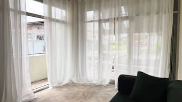 A room with a green sofa, white curtains, and a view of a balcony with a metal railing.