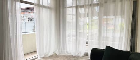 Una habitación con sofá verde, cortinas blancas y vistas a un balcón con barandilla de metal.