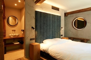 Ein Schlafzimmer mit einem Bett, einem runden Spiegel, einem Nachttisch aus Holz, einer eingebauten Ablage mit einem Waschbecken und einem Kopfteil mit vertikalen Paneelen.