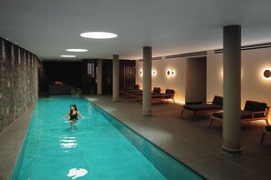 An indoor swimming pool with a person swimming, lounge chairs, and recessed ceiling lights. - Magma Resort Santorini in of the Unbound Collection by Hyatt (Santorini)