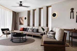Un salon moderne avec un canapé, une table basse et des fauteuils. On y trouve un ventilateur de plafond, un luminaire fixé au mur et une fenêtre avec des rideaux.