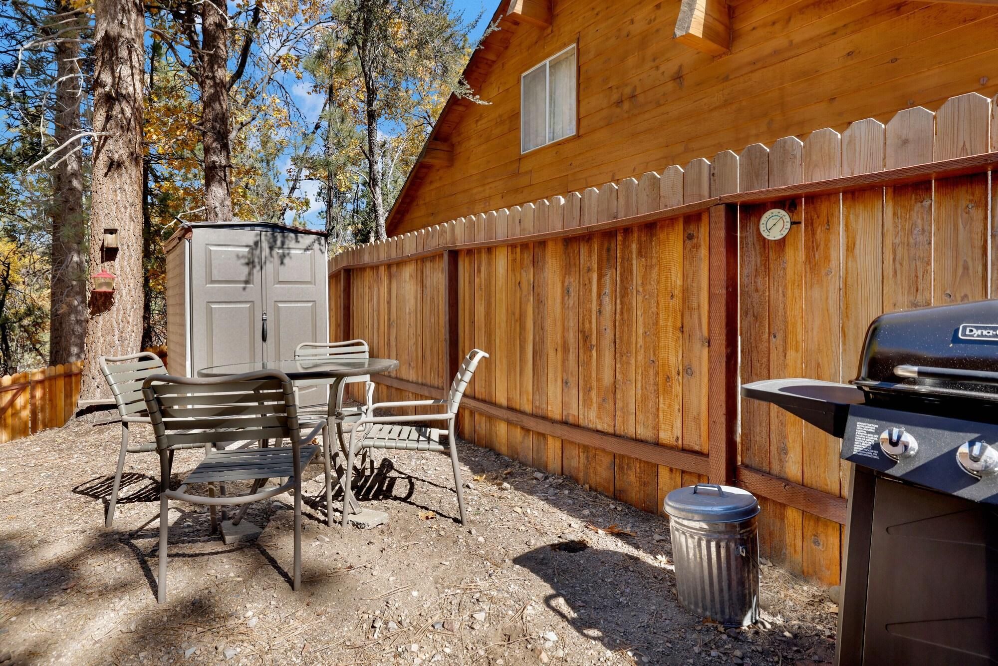 A wooden cabin with a barbecue grill, a table with chairs, and a trash can.
