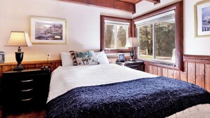 Un dormitorio con una cama, dos mesitas de noche, una lámpara, un cuadro enmarcado y una ventana con vista a árboles.