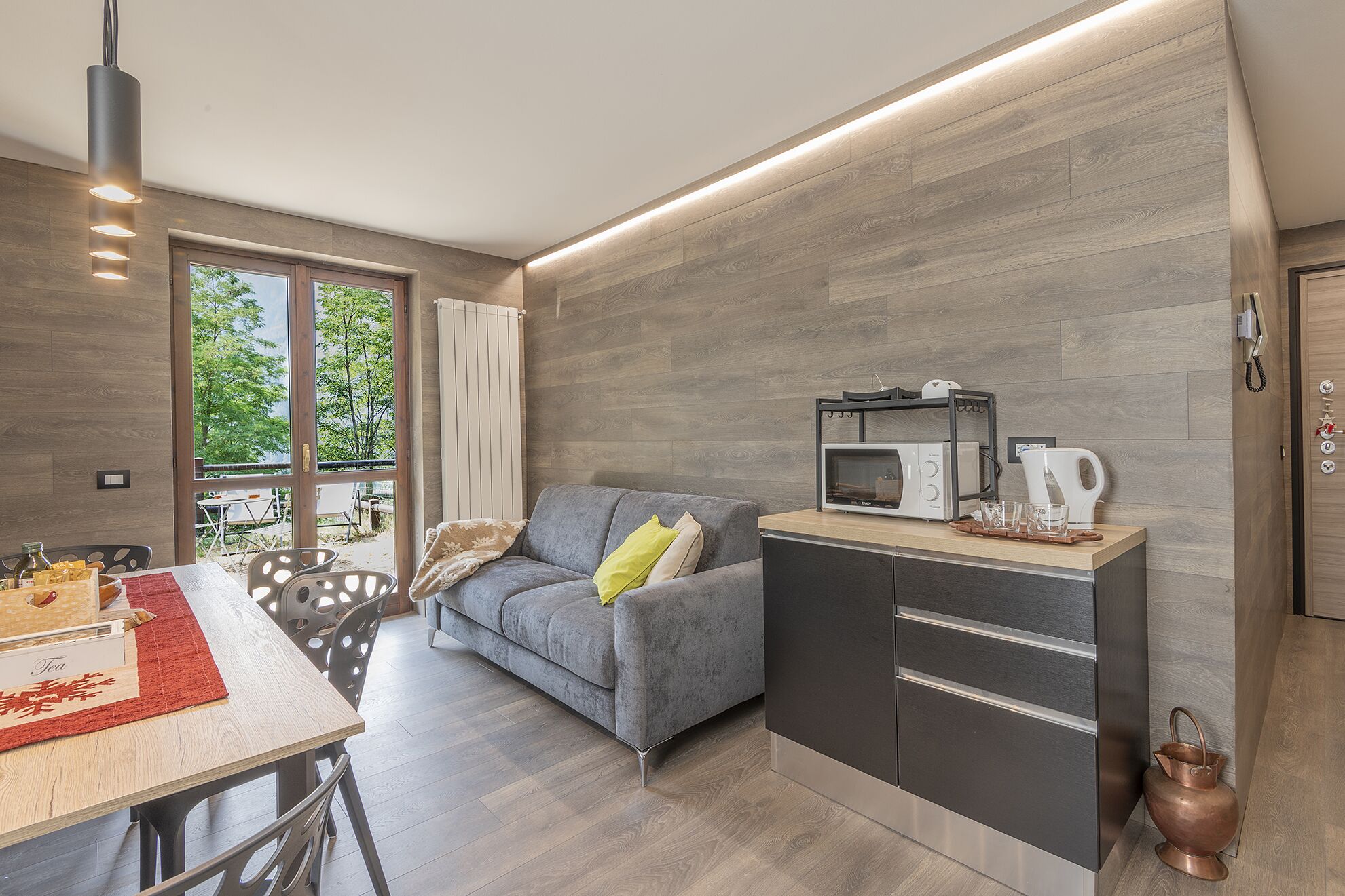 A modern kitchen with a grey sofa, a dining table with chairs, and a view of greenery outside.