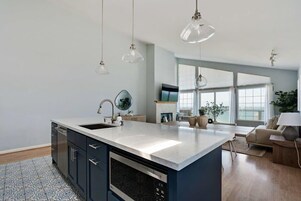 Cozinha moderna com uma ilha grande, iluminação pendente e vista para uma sala de estar.